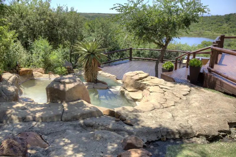 A rock pool cut into the hillside beside a timber deck, with an aloe growing at its edge and the bush and a dam stretching away below.