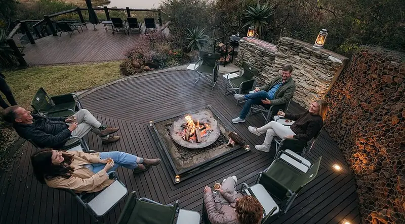 Six guests in canvas chairs around a square fire pit on a timber deck at dusk, with lanterns hung along the stone wall behind them.