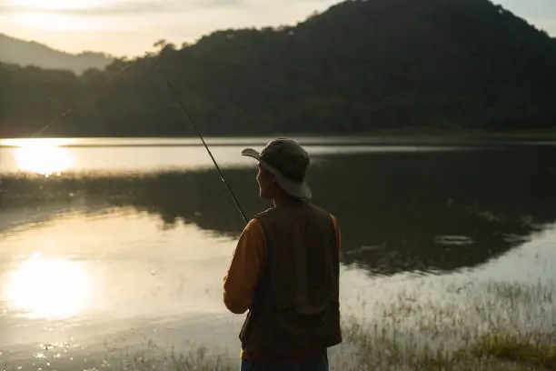 A lone angler in a wide-brimmed hat casting into a still lake at dawn, with wooded hills rising behind.