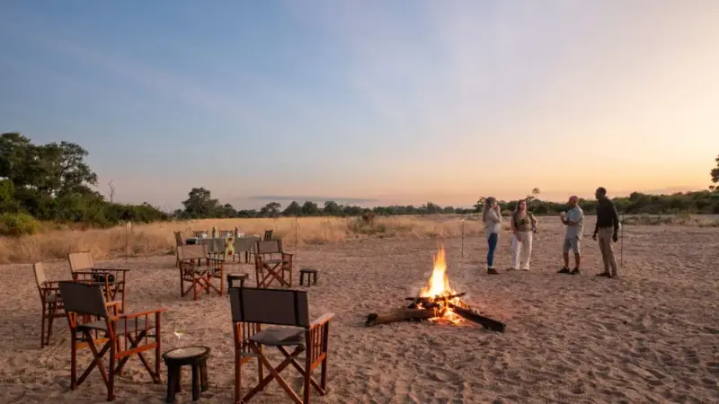 Camp chairs and a laid table set out on a dry riverbed at dusk, a fire burning in the foreground and four people standing talking beyond it.