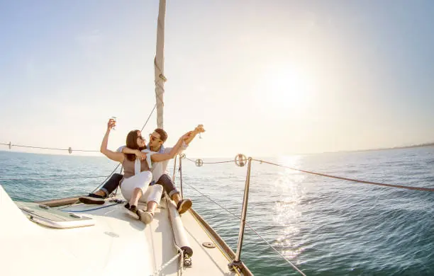 A couple sitting close together on the foredeck of a sailing boat at sunset, raising a glass each, with calm open water stretching to the horizon behind them.
