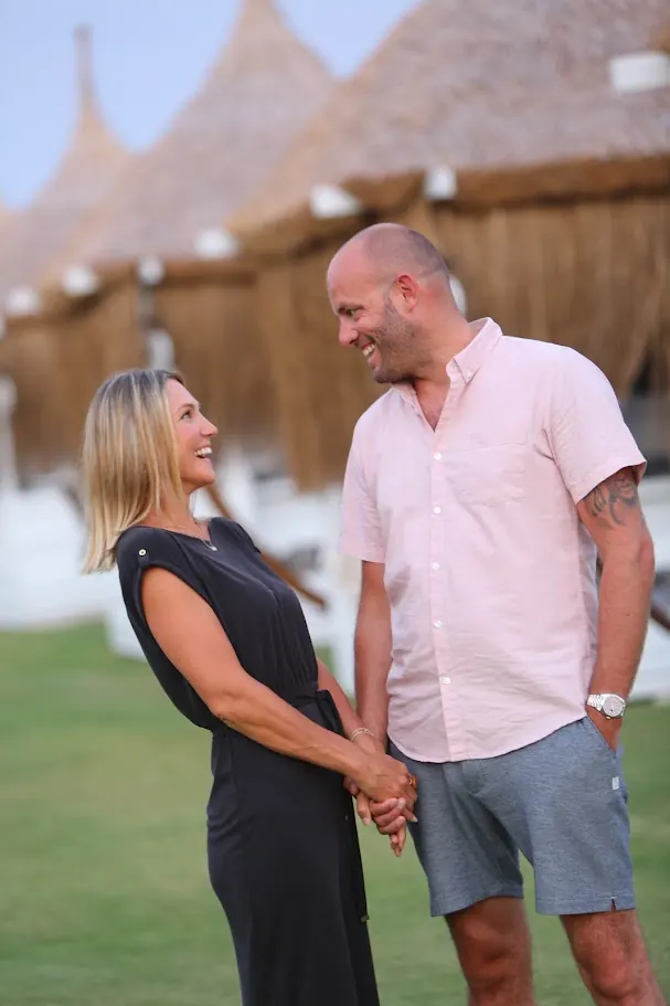 Stacey and Ollie laughing together on a lawn in front of a row of thatched beach cabanas at dusk.