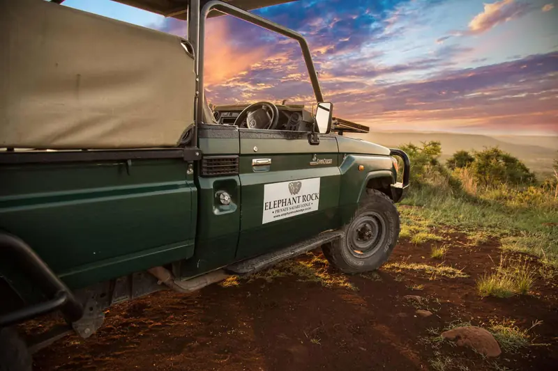 An open-sided safari vehicle parked on a ridge at dawn, with the bush and low hills stretching away under a streaked pink sky.