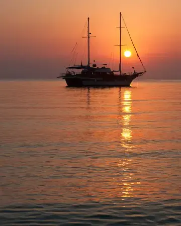 A gulet at anchor silhouetted against a low orange sun, its reflection running across still water.