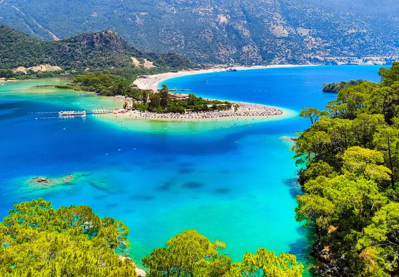 The lagoon at Ölüdeniz seen from above, a curving spit of pale sand separating shallow turquoise water from the deeper blue of the bay, with pine-covered mountains behind.