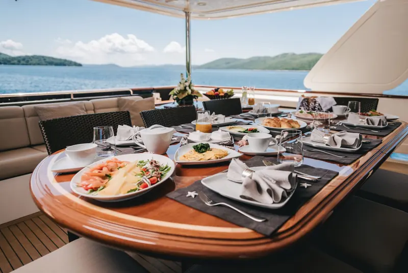 A long table laid for breakfast on a yacht's shaded aft deck, plates of eggs, pastries and cured fish set out with coffee cups and glasses, and calm sea and low green islands beyond.